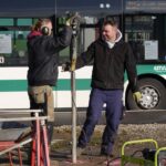 Zwei Mitarbeiter führen eine Kleinbohrung am Straßenrand durch. Das Gestänge ist so groß wie die beiden Männer. Im Vordergrund stehen ein Stromaggregat, eine Schubkarre und Aufstellböcke zum Ablegen der Sonden. Im Hintergrund fährt ein Bus vorbei