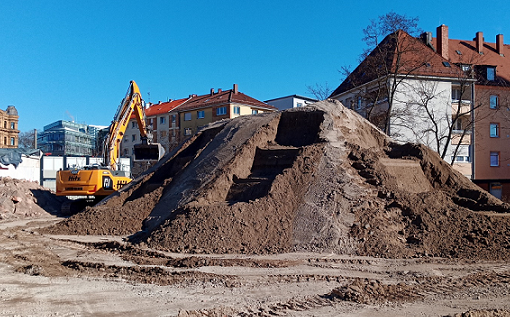 Ein Bagger türmt einen großen Erdhaufen auf einem Grundstück im städtischen Raum auf. Ins Haufwerk sind Stufen ausgehoben, um die optimale Probenahme zu ermöglichen.