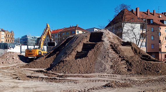 Ein Bagger türmt einen großen Erdhaufen auf einem Grundstück im städtischen Raum auf. Ins Haufwerk sind Stufen ausgehoben, um die optimale Probenahme zu ermöglichen.